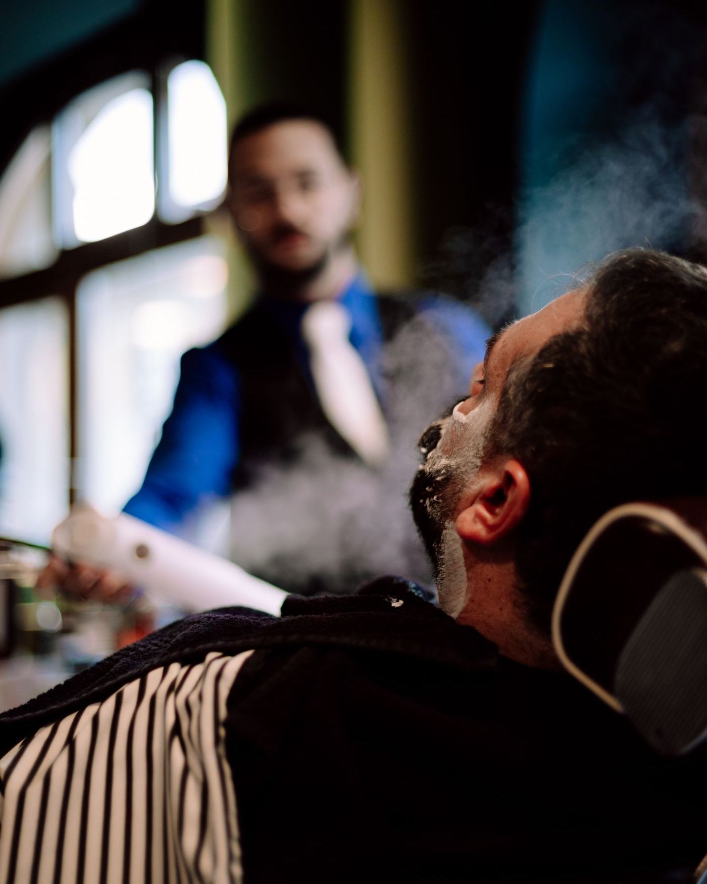 A barber in a vest and tie readies a hot-towel shave for a client reclined in a leather chair, arched windows behind them