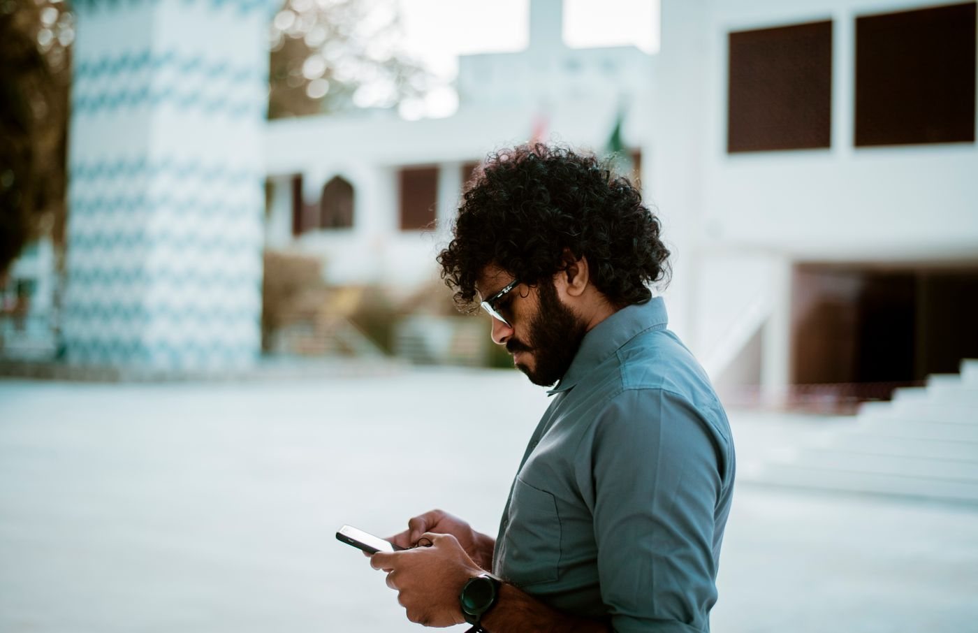 A man with curly hair and sunglasses stands outside in the sun, looking down at his phone