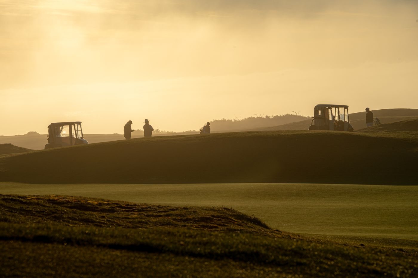 Small figures of golfers and carts on a rolling course in golden evening haze