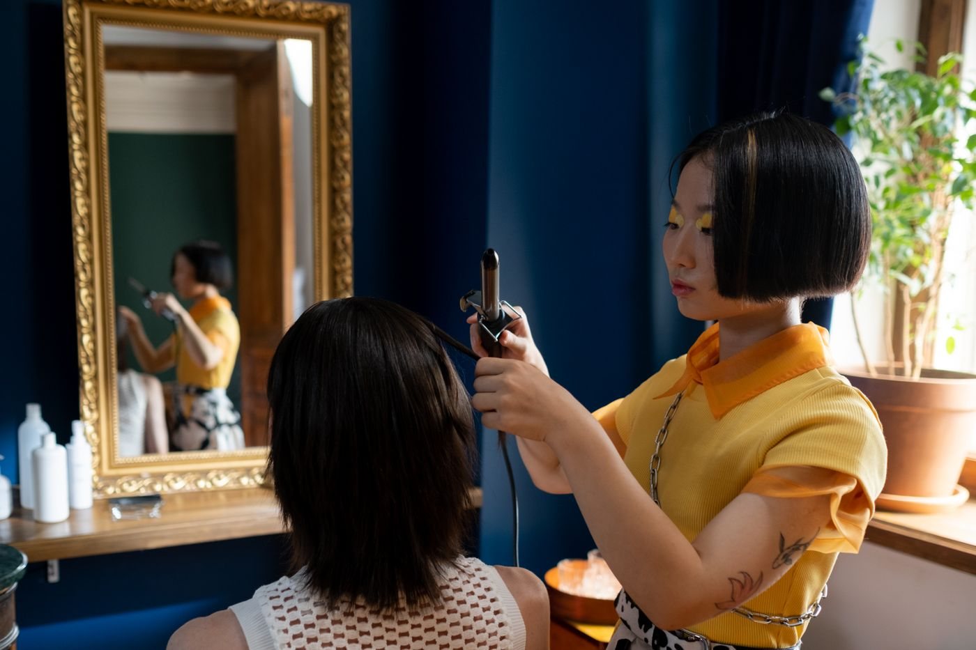 A stylist with a sharp bob curls a client's hair in a city salon with deep blue walls, a gilt mirror and plants by the window