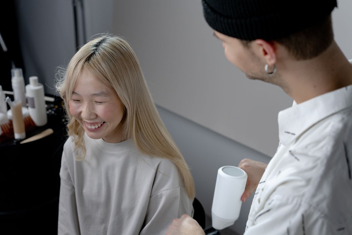 A young woman with long blonde hair laughs as her stylist, dryer in hand, finishes her blow-dry