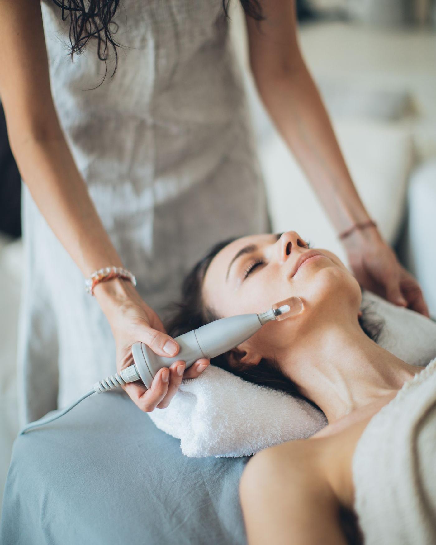 A woman relaxes with her eyes closed while an aesthetician moves a skin-treatment wand along her jaw